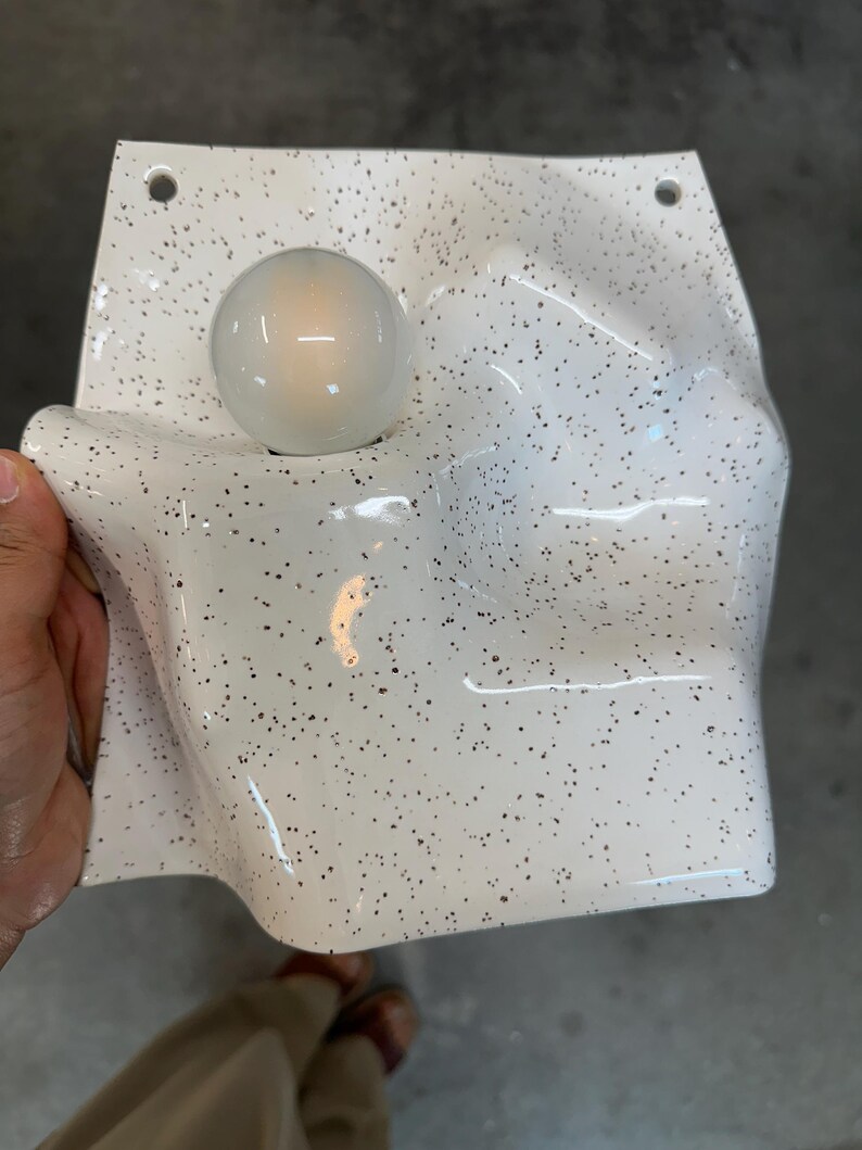 May include: A white wall sconce with a speckled black pattern and a glossy finish. The fixture features an irregular, undulating surface with a single round, glowing light bulb integrated into the left side and two mounting holes at the top corners.