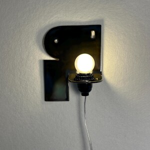 May include: A wall-mounted sconce featuring a glossy black ceramic backplate with an irregular, curved shape. A single round, glowing white light bulb is mounted on a black socket extending from the plate, with a thin, clear power cord hanging below.
