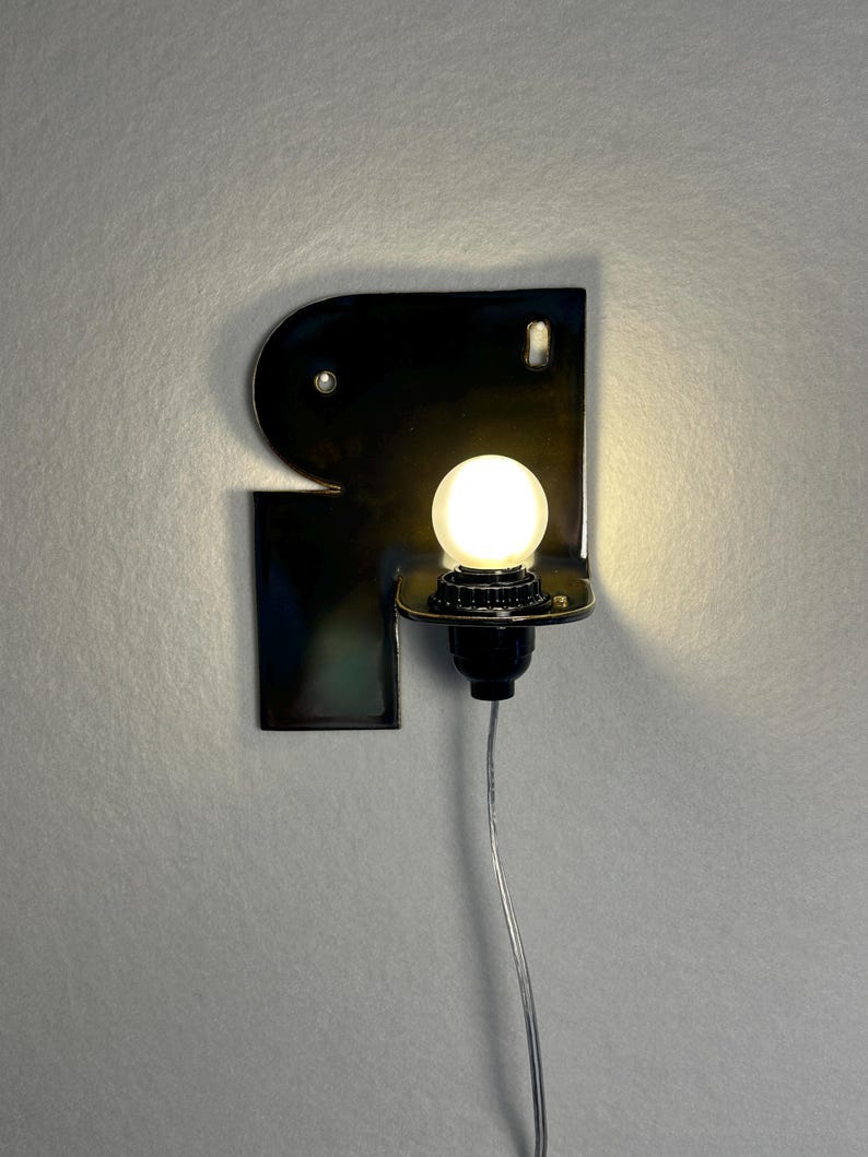 May include: A wall-mounted sconce featuring a glossy black ceramic backplate with an irregular, curved shape. A single round, glowing white light bulb is mounted on a black socket extending from the plate, with a thin, clear power cord hanging below.