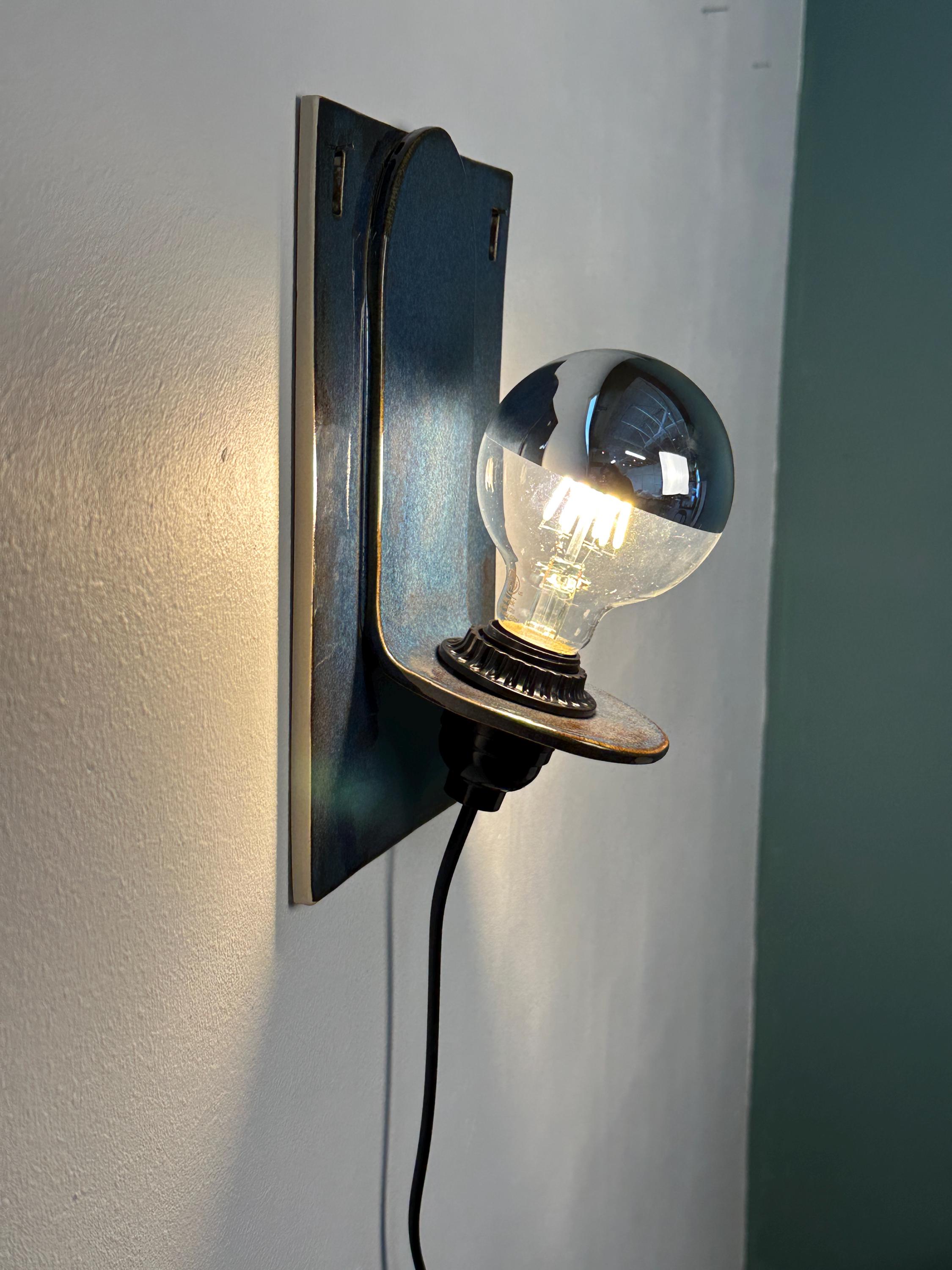 May include: A wall-mounted light fixture featuring a rectangular blue ceramic backplate with a curved shelf. A black socket holds a round, clear glass light bulb with a silver-dipped top and visible internal filaments, connected by a black cord.