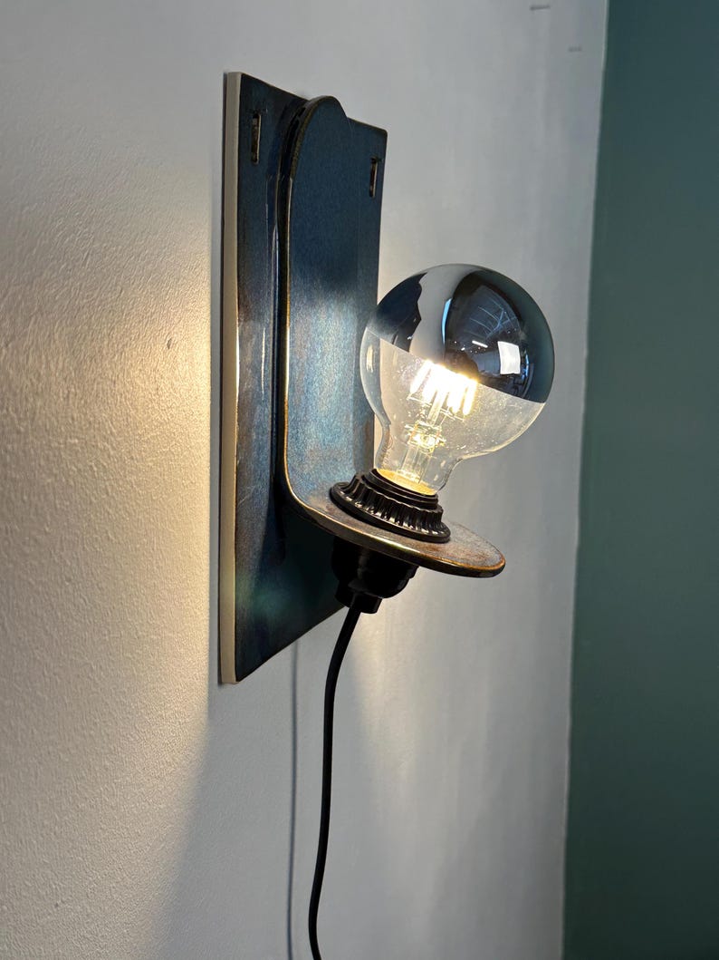 May include: A wall-mounted light fixture featuring a rectangular blue ceramic backplate with a curved shelf. A black socket holds a round, clear glass light bulb with a silver-dipped top and visible internal filaments, connected by a black cord.