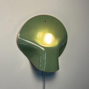 May include: A sage green ceramic wall sconce with a smooth, glossy finish and an organic, curved shape. The fixture features a central circular opening holding a glowing, frosted white light bulb and a small hole at the top for mounting.