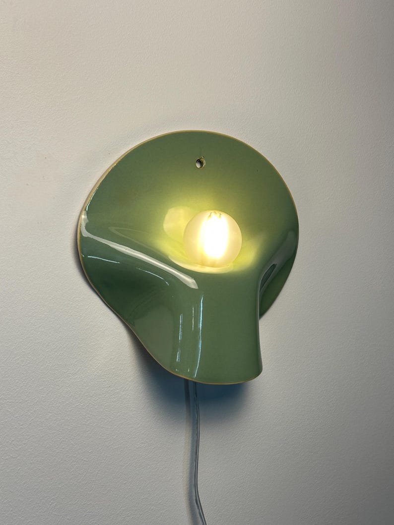 May include: A sage green ceramic wall sconce with a smooth, glossy finish and an organic, curved shape. The fixture features a central circular opening holding a glowing, frosted white light bulb and a small hole at the top for mounting.