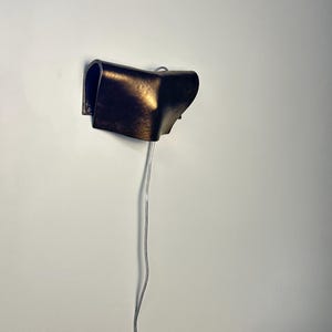 May include: A wall-mounted lamp featuring a curved, dark bronze-toned metal shade with a hammered texture. The fixture has a folded, sculptural form and includes a thin, light-coloured electrical cord extending downward from the base of the unit.
