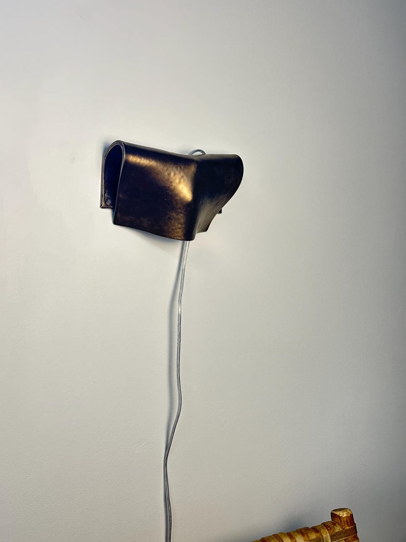 May include: A wall-mounted lamp featuring a curved, dark bronze-toned metal shade with a hammered texture. The fixture has a folded, sculptural form and includes a thin, light-coloured electrical cord extending downward from the base of the unit.