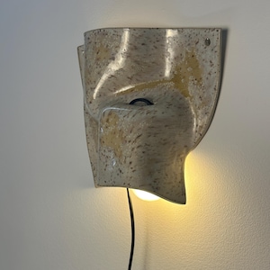 May include: A wall-mounted ceramic sconce with a speckled cream and tan finish. The fixture features a curved, organic shape with a central opening that emits a warm, glowing light from an internal bulb. A thin black power cord hangs downward.