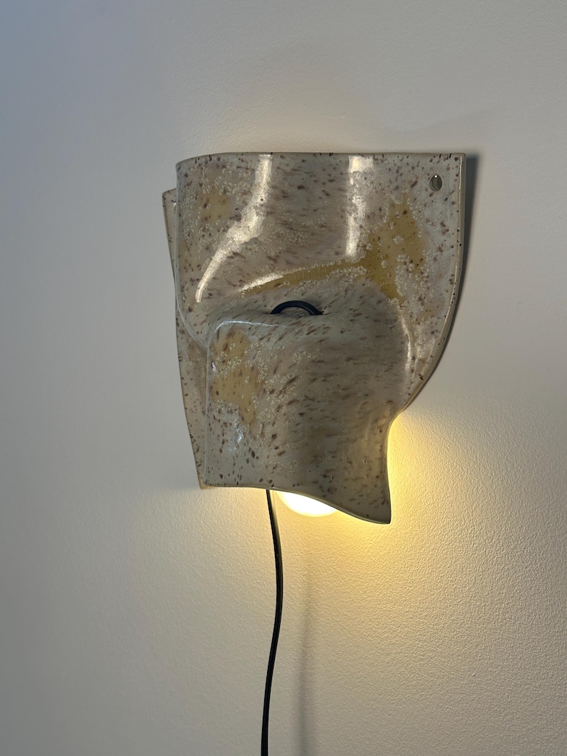 May include: A wall-mounted ceramic sconce with a speckled cream and tan finish. The fixture features a curved, organic shape with a central opening that emits a warm, glowing light from an internal bulb. A thin black power cord hangs downward.