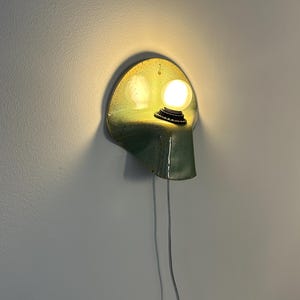 Puede incluir: Un aplique de pared con una pantalla de cer&aacute;mica curva, brillante y moteada en tonos verde y chartreuse. Una bombilla blanca redonda e iluminada est&aacute; montada en el lado derecho, con un cable de alimentaci&oacute;n blanco y fino que cuelga hacia abajo.