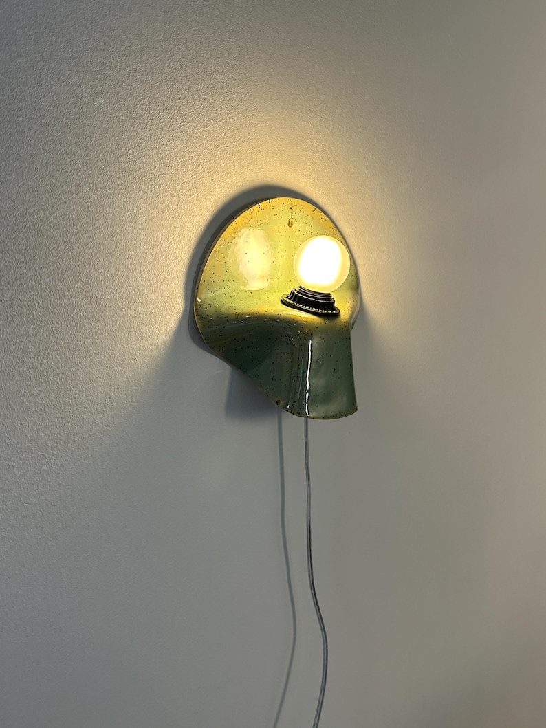 Puede incluir: Un aplique de pared con una pantalla de cer&aacute;mica curva, brillante y moteada en tonos verde y chartreuse. Una bombilla blanca redonda e iluminada est&aacute; montada en el lado derecho, con un cable de alimentaci&oacute;n blanco y fino que cuelga hacia abajo.