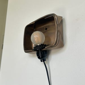 May include: A bronze-colored wall sconce with a light bulb. The sconce has a rectangular shape with a curved top and is mounted on a white wall. The light bulb is round and has a black base with a black cord.