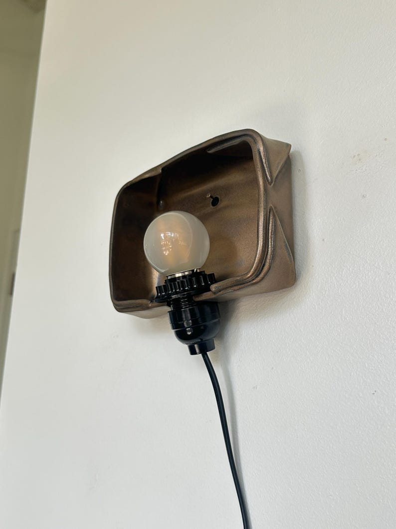 May include: A bronze-colored wall sconce with a light bulb. The sconce has a rectangular shape with a curved top and is mounted on a white wall. The light bulb is round and has a black base with a black cord.