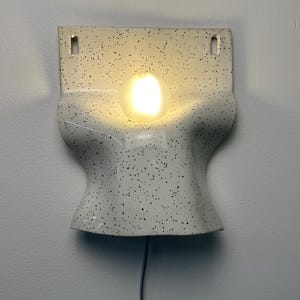 Może przedstawiać: Kinkiet ceramiczny z kremowym szkliwem i czarnymi kropkami. Oprawa ma zakrzywiony, formowany kształt z dwoma prostokątnymi otworami montażowymi u g&oacute;ry oraz centralnym okrągłym otworem, w kt&oacute;rym znajduje się świecąca żar&oacute;wka.
