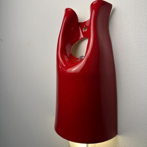 May include: A glossy red wall-mounted light fixture with a smooth, curved, sculptural form. The piece features an open cutout near the top for mounting and a single light bulb emitting a warm glow from the bottom, connected by a clear power cord.
