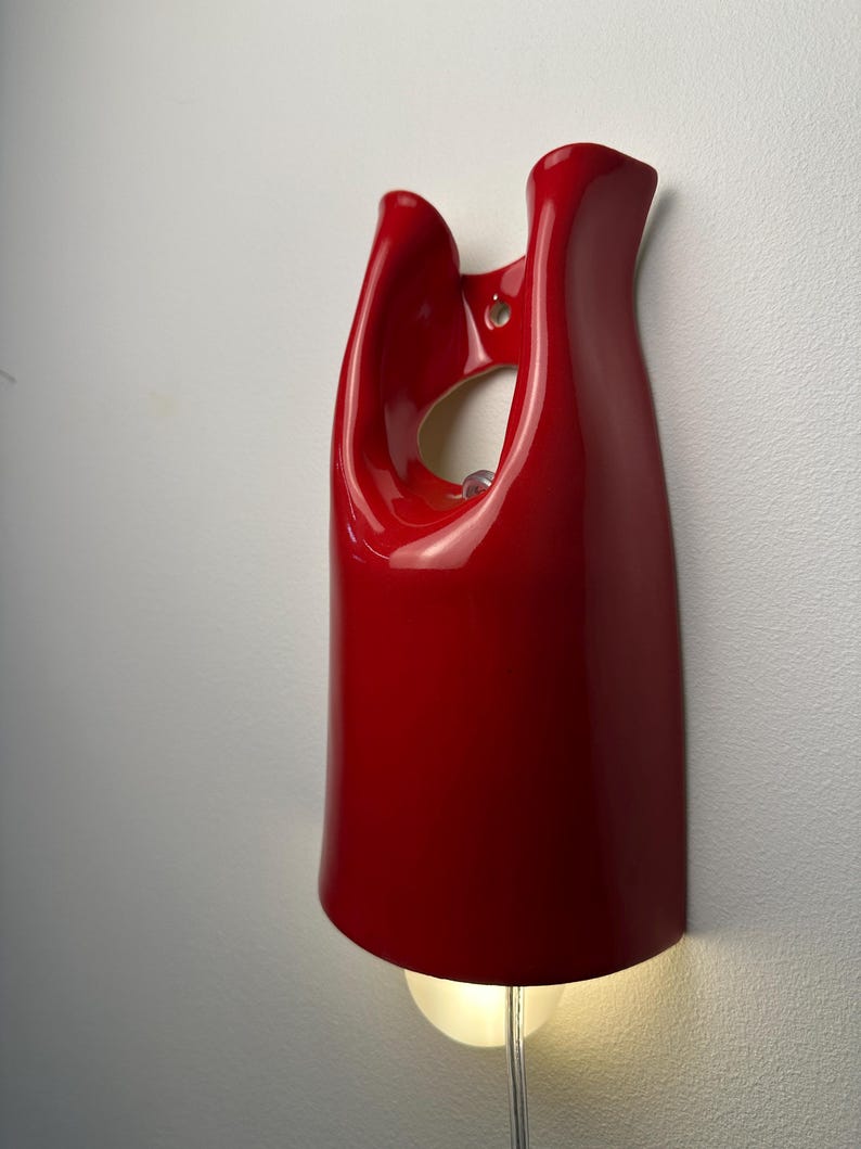 May include: A glossy red wall-mounted light fixture with a smooth, curved, sculptural form. The piece features an open cutout near the top for mounting and a single light bulb emitting a warm glow from the bottom, connected by a clear power cord.