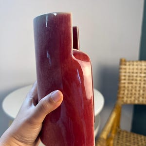May include: A tall, cylindrical ceramic vase with a deep, glossy burgundy red glaze. The vase features a unique stepped top design with one side significantly higher than the other, creating a distinct architectural silhouette with smooth curves.