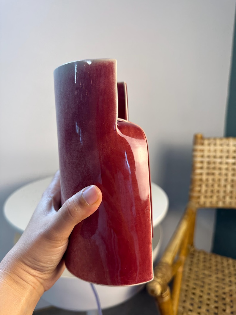 May include: A tall, cylindrical ceramic vase with a deep, glossy burgundy red glaze. The vase features a unique stepped top design with one side significantly higher than the other, creating a distinct architectural silhouette with smooth curves.