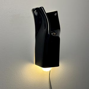 May include: A glossy black ceramic wall sconce with a curved, sculptural form. The fixture features two visible mounting holes at the top and emits a warm, downward-facing light from a single bulb. A thin power cord extends from the bottom.