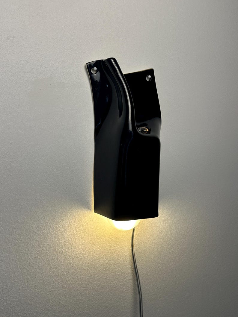 May include: A glossy black ceramic wall sconce with a curved, sculptural form. The fixture features two visible mounting holes at the top and emits a warm, downward-facing light from a single bulb. A thin power cord extends from the bottom.