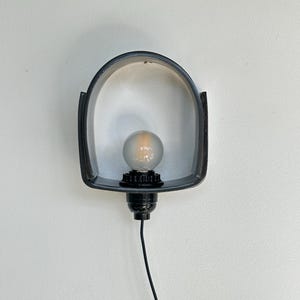 Puede incluir: Una l&aacute;mpara de pared negra con un marco curvo en forma de arco. Cuenta con una bombilla redonda y un cable negro. La luz est&aacute; montada en una pared blanca. Dise&ntilde;o sencillo y moderno.
