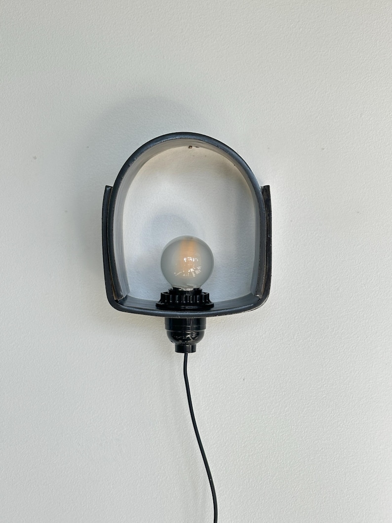 Puede incluir: Una l&aacute;mpara de pared negra con un marco curvo en forma de arco. Cuenta con una bombilla redonda y un cable negro. La luz est&aacute; montada en una pared blanca. Dise&ntilde;o sencillo y moderno.