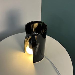 May include: A table lamp featuring a glossy black curved cylindrical shade that partially wraps around a single glowing light bulb. The lamp includes a black socket and a thin white power cord extending from the side of the structure.