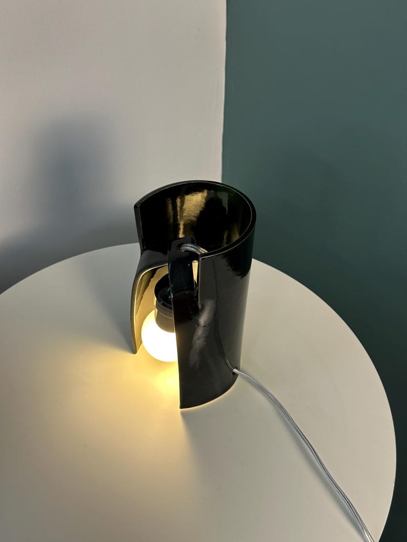 May include: A table lamp featuring a glossy black curved cylindrical shade that partially wraps around a single glowing light bulb. The lamp includes a black socket and a thin white power cord extending from the side of the structure.