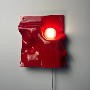 May include: A glossy red wall lamp with a wavy, undulating surface and a circular cutout holding a glowing white spherical light bulb. The fixture features two small holes at the top corners for mounting and a thin gray power cord extending downward.