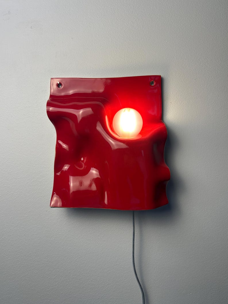 May include: A glossy red wall lamp with a wavy, undulating surface and a circular cutout holding a glowing white spherical light bulb. The fixture features two small holes at the top corners for mounting and a thin gray power cord extending downward.