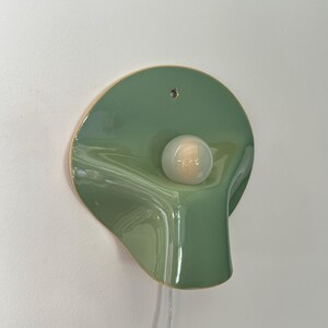 May include: A wall-mounted ceramic light fixture with a glossy sage green finish. The piece features an organic, curved, petal-like shape with a central circular opening holding a small, round, frosted white light bulb and a clear power cord.