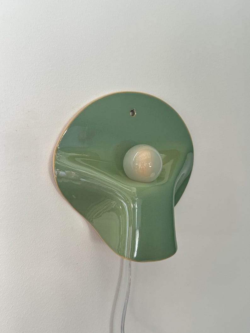May include: A wall-mounted ceramic light fixture with a glossy sage green finish. The piece features an organic, curved, petal-like shape with a central circular opening holding a small, round, frosted white light bulb and a clear power cord.