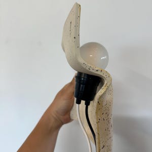 May include: A wall-mounted light fixture featuring a curved, off-white ceramic shade with a speckled pattern. The fixture includes a black socket holding a round light bulb, with two exposed electrical wires, one white and one black, extending downward.