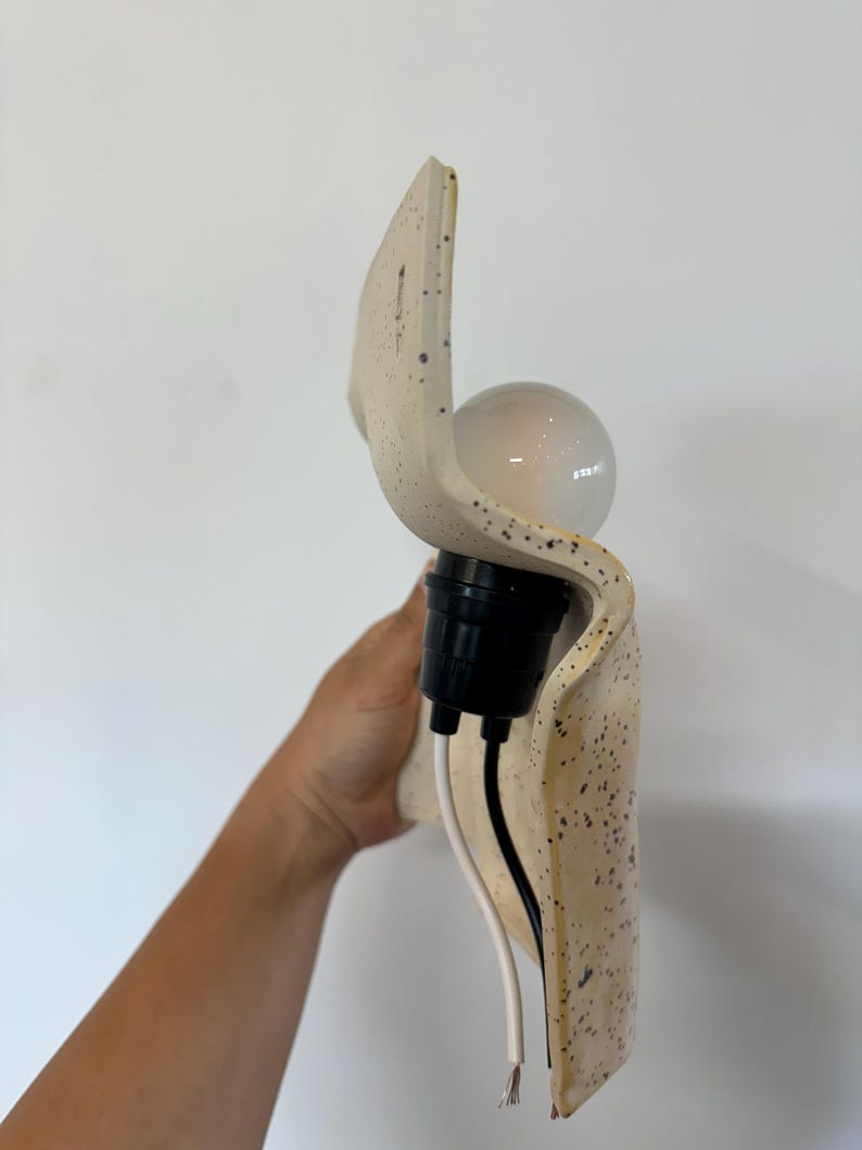 May include: A wall-mounted light fixture featuring a curved, off-white ceramic shade with a speckled pattern. The fixture includes a black socket holding a round light bulb, with two exposed electrical wires, one white and one black, extending downward.