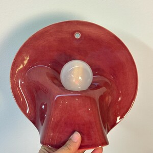May include: A glossy, deep red ceramic wall sconce featuring a wide, undulating, shell-like shade with a central circular opening. A single, spherical, frosted white light bulb is positioned in the centre of the fixture, which includes a small hole at the top for mounting.