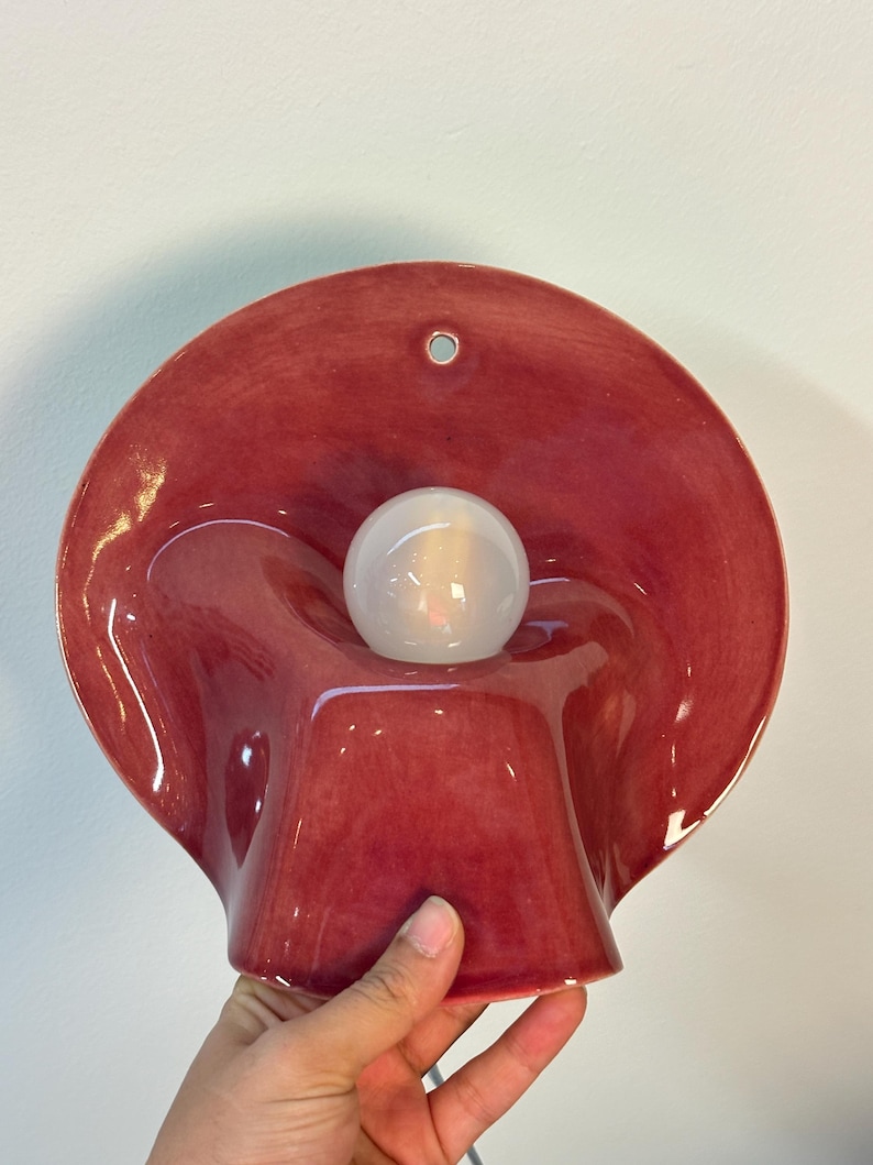 May include: A glossy, deep red ceramic wall sconce featuring a wide, undulating, shell-like shade with a central circular opening. A single, spherical, frosted white light bulb is positioned in the centre of the fixture, which includes a small hole at the top for mounting.