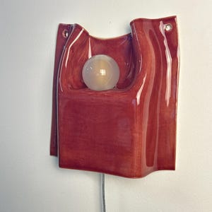 Puede incluir: Un aplique de pared de cer&aacute;mica con un acabado rojo intenso brillante y una forma org&aacute;nica ondulada. La pieza incluye un hueco circular central que sostiene una bombilla de globo blanca esmerilada y dos orificios superiores.