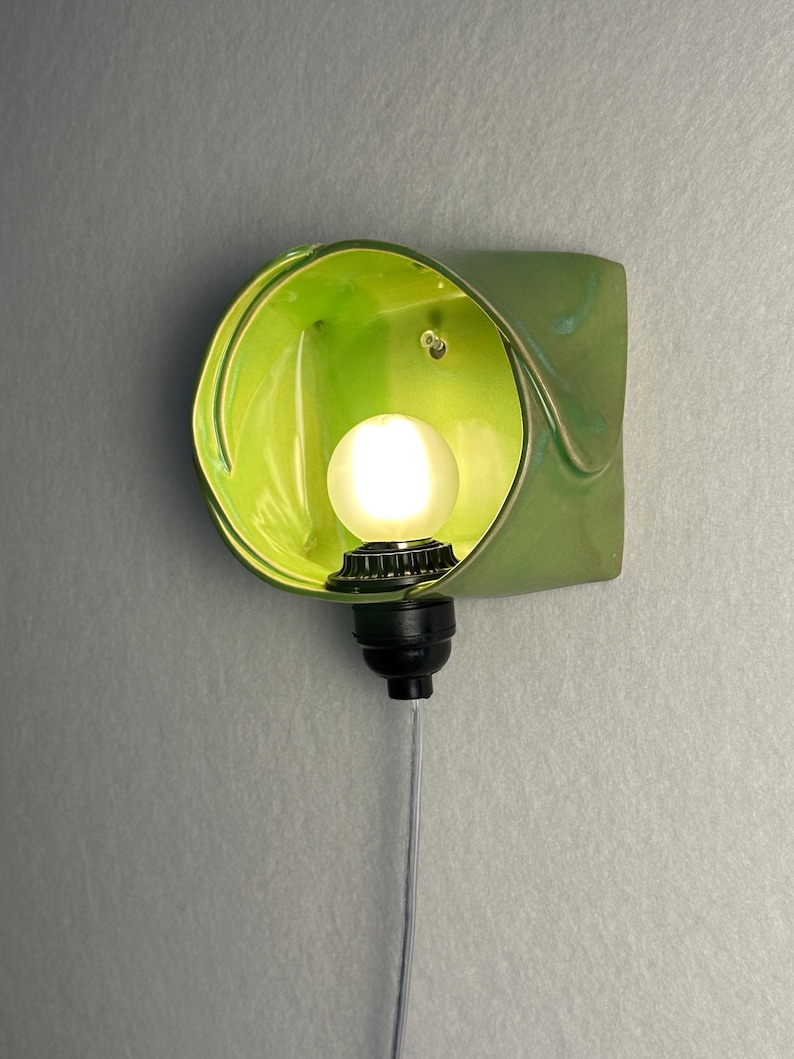 May include: A wall-mounted light fixture featuring a glossy, lime green ceramic shade with an organic, folded shape. A small, round, frosted white light bulb is centered within the shade, attached to a black socket with a thin, light-colored cord.