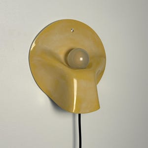 May include: A wall-mounted lamp featuring a circular, glossy yellow ceramic shade with a fluid, organic shape. A single, spherical, frosted white light bulb is centered within the recessed portion of the shade. A black power cord extends downward.
