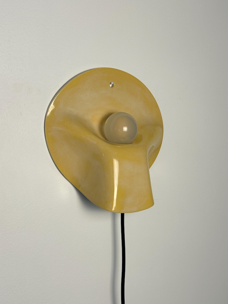 May include: A wall-mounted lamp featuring a circular, glossy yellow ceramic shade with a fluid, organic shape. A single, spherical, frosted white light bulb is centered within the recessed portion of the shade. A black power cord extends downward.