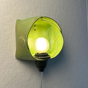 May include: A wall-mounted light fixture featuring a glossy, lime green, organic-shaped ceramic shade. A small, round, frosted white light bulb is centered within the shade, attached to a black socket base with a thin, vertical power cord.