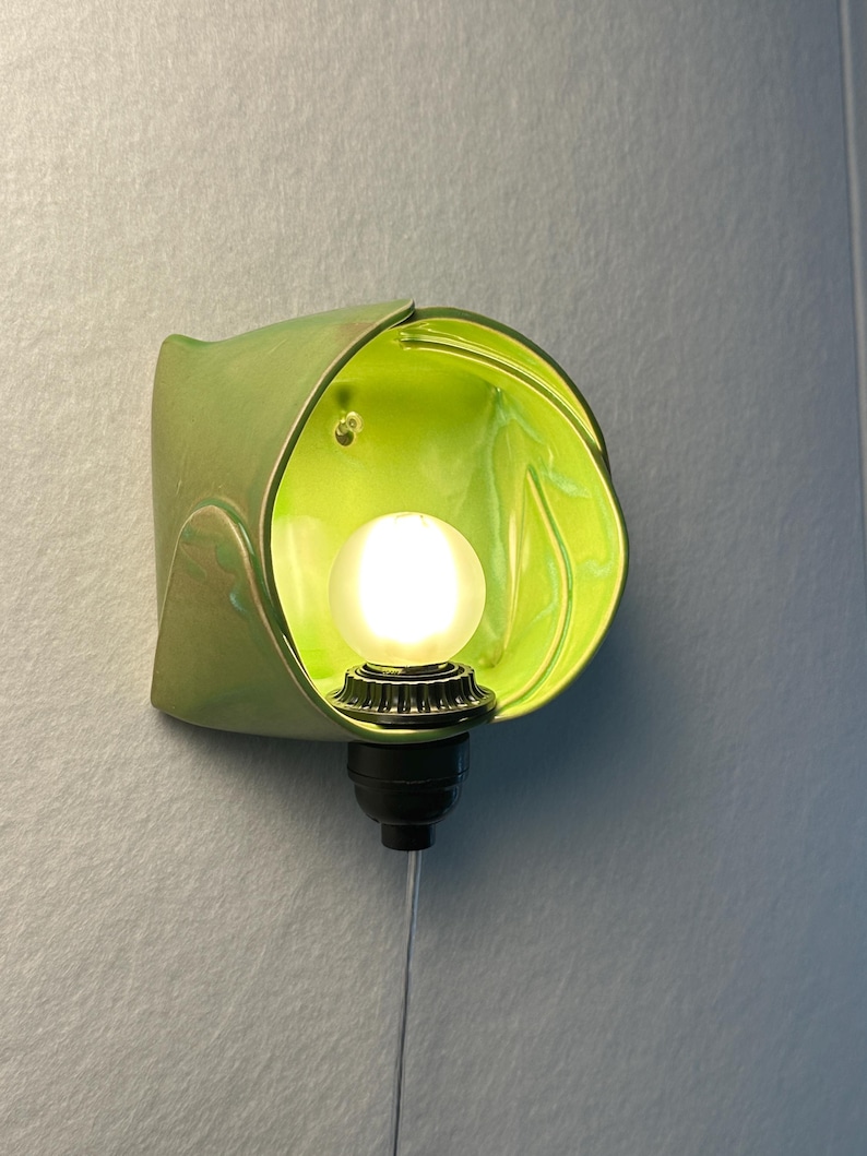 May include: A wall-mounted light fixture featuring a glossy, lime green, organic-shaped ceramic shade. A small, round, frosted white light bulb is centered within the shade, attached to a black socket base with a thin, vertical power cord.