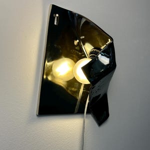 Peut inclure: Une applique murale compos&eacute;e d'un abat-jour en c&eacute;ramique noire brillante &agrave; la forme irr&eacute;guli&egrave;re et repli&eacute;e. Le luminaire comprend une ampoule expos&eacute;e diffusant une lumi&egrave;re chaude qui se refl&egrave;te sur la surface sombre et brillante de la c&eacute;ramique.