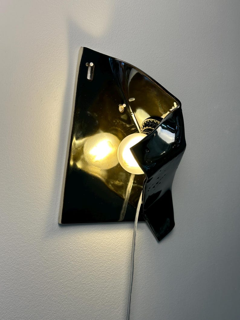 Peut inclure: Une applique murale compos&eacute;e d'un abat-jour en c&eacute;ramique noire brillante &agrave; la forme irr&eacute;guli&egrave;re et repli&eacute;e. Le luminaire comprend une ampoule expos&eacute;e diffusant une lumi&egrave;re chaude qui se refl&egrave;te sur la surface sombre et brillante de la c&eacute;ramique.