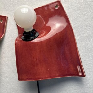 Puede incluir: Una l&aacute;mpara de pared con una placa de cer&aacute;mica de color rojo intenso brillante y forma irregular y curva. Una bombilla esf&eacute;rica blanca est&aacute; montada en el lado izquierdo en un casquillo negro. La palabra TAJADDOD est&aacute; grabada en la esquina inferior derecha.