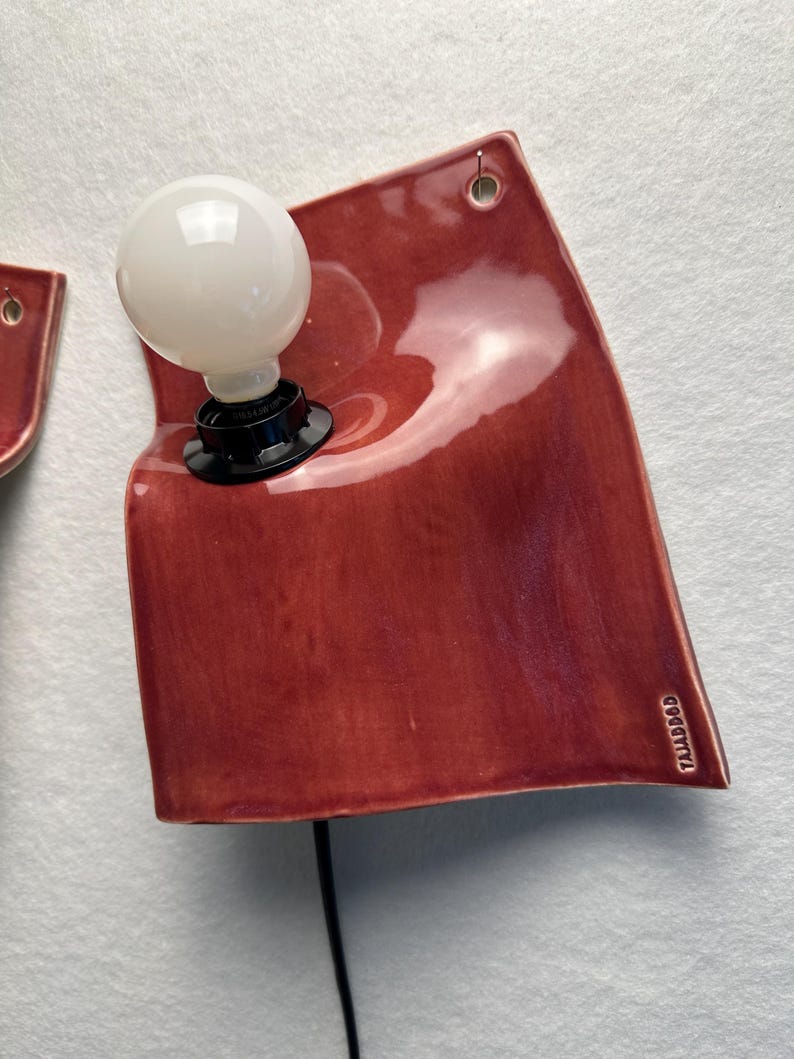 Puede incluir: Una l&aacute;mpara de pared con una placa de cer&aacute;mica de color rojo intenso brillante y forma irregular y curva. Una bombilla esf&eacute;rica blanca est&aacute; montada en el lado izquierdo en un casquillo negro. La palabra TAJADDOD est&aacute; grabada en la esquina inferior derecha.