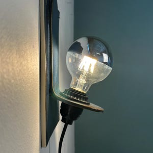 May include: A wall-mounted lamp featuring a dark, flat vertical backplate and a protruding circular support holding a single clear glass light bulb with a visible filament. A black cord extends downward from the black socket base.