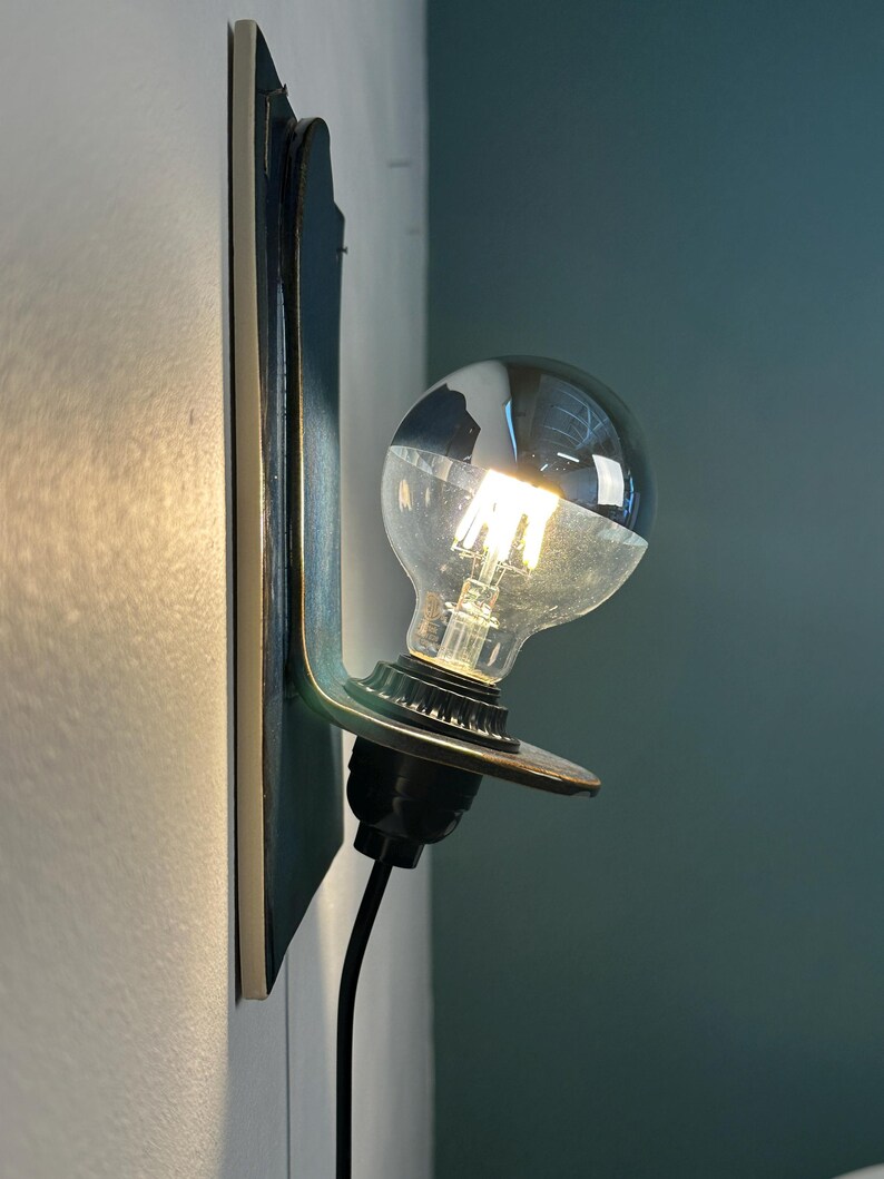 May include: A wall-mounted lamp featuring a dark, flat vertical backplate and a protruding circular support holding a single clear glass light bulb with a visible filament. A black cord extends downward from the black socket base.
