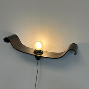 May include: A modern wall sconce with a curved, dark brown metal design. The sconce features a single, round light bulb in the center, emitting a warm glow. A gray power cord extends downward. The fixture is mounted on a white wall.