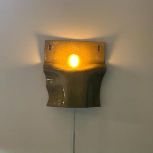 May include: A wall-mounted ceramic sconce with a speckled olive-green glaze and a curved, sculptural form. The fixture emits a warm, glowing light from a central bulb, casting soft shadows against the wall. A thin white power cord hangs below.