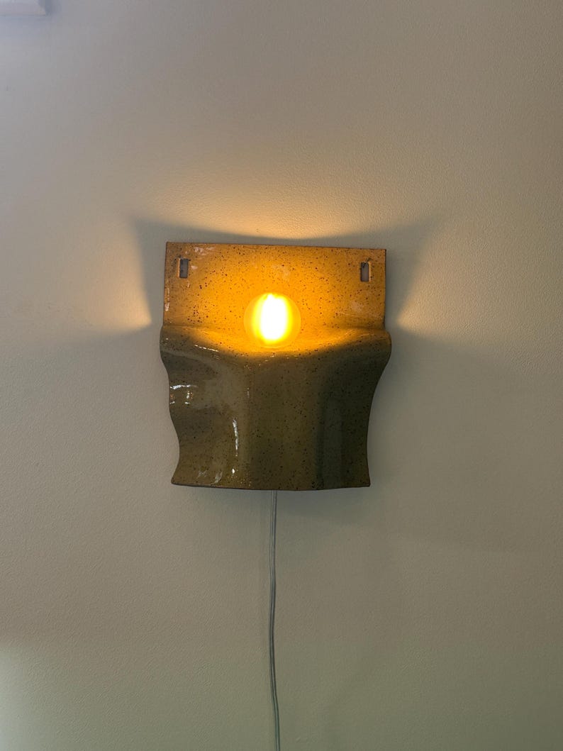May include: A wall-mounted ceramic sconce with a speckled olive-green glaze and a curved, sculptural form. The fixture emits a warm, glowing light from a central bulb, casting soft shadows against the wall. A thin white power cord hangs below.
