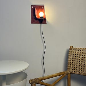 May include: A wall-mounted lamp featuring a rectangular, dark red ceramic backplate with a curved, bowl-like center. A black socket holds a glowing, round light bulb. A thin black power cord hangs vertically from the base of the light fixture.